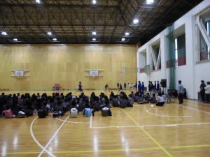 体育館の前方で部活を紹介する生徒が披露し、その様子を床に座り見ている様子の写真