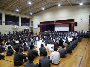 体育館の舞台前に生徒が並び、床に座っている生徒が見ている様子を後ろから写した写真