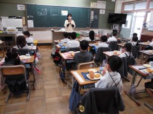 自席のランチョンマットの上に給食が置かれ、教卓の前で先生が手を合わせ児童も手を合わせている写真