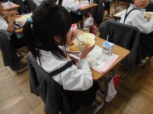 自席で女の子がスープを持ちスプーンで食べてる様子の写真