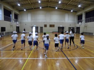 体育館のステージ側から、反対側の出入口に向かい、白い帽子や体操服、青い短パンを着用した10名ほどの生徒達が、一生懸命走っている様子を背後から撮影した写真