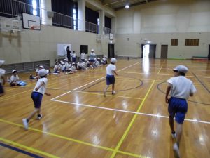 壁沿いに同級生が見守る中、ステージ側から体育館後方に向かい3名の生徒達が走っている様子を背後から撮影した写真