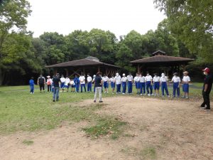 周りを緑色の木々に囲まれた信太山青少年活動センターの広場で、横一列になり話を聞く生徒達と生徒達と向かい合わせで話をする先生方の写真