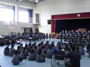 体育館の舞台の前に座った生徒、左側に立っている生徒、右側に床に座っている生徒6年生を笑顔で送る会の様子を写した写真