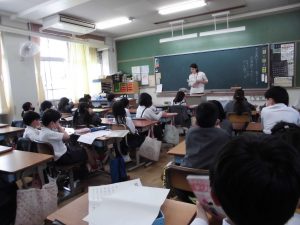 自席の上に真新しい教科書やプリントを広げ、教卓にいる先生の話を静かに話を聞く生徒達を背後から撮影した写真