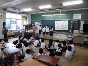 白い大きな模造紙を貼り付けた黒板前で、校区体験について発表している4名の生徒達と、向かい合わせで膝を抱えるように座りながら熱心に話を聞く生徒を背後から撮影した写真