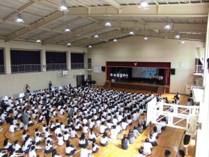 体育館の壇上に設置されたスクリーンを使い発表している生徒達と、熱心に話を聞く生徒達を2階のキャットウォークから撮影した写真