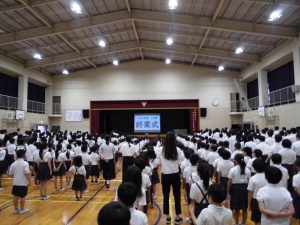 白く清潔感のある広い体育館内で、全校生徒達が立ち整列し、壇上の終業式と表示されたスクリーンを注目している様子を左背後から撮影した写真