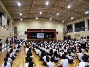 奥行きのあるきれいな体育館内で、整列した全校生徒達が三角座りで壇上のスクリーンを見ている様子を左背後から撮影した写真