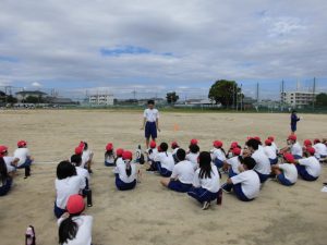 運動場で一人の生徒が立って説明しているのを座って生徒たちが話を聞いている様子の写真
