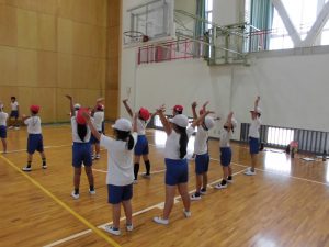 体育館で等間隔に生徒たちが3列ほどに並び、両手を上に挙げたりして体を動かしている写真