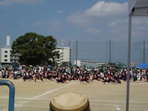 学校の運動場でハチマキや衣装を着た生徒たちが演舞をしている様子を遠目で撮影した写真
