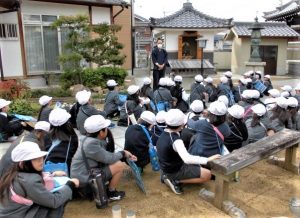 白い帽子を被った児童達が、西稍寺住職の話を境内の地面に座り話しを聞いてる様子の写真