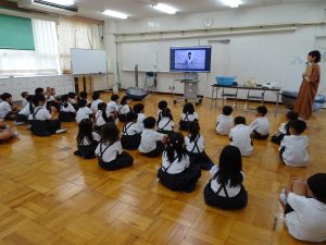教室内に設置されたテレビモニター画面に映し出され、膝を抱えるように座りながら熱心に話を聞く生徒達を背後から撮影した写真
