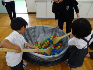 給食を作る際に使用する、大きな鍋や木べらを使い、食材に見立てたカラーボールを大きな木べらで一生懸命かき混ぜる体験をしている男女2名の生徒の写真