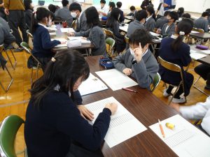 教室で長い机に向かい合いで3人が座り、1人が話をして、後の2人が話を聞いたりメモをしてる様子の写真