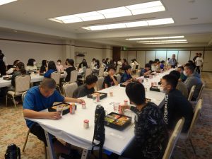 宴会場のような広さがある室内で2列に並んだ長テーブルに向かい合うように生徒たちが椅子に座りお弁当を食べている写真