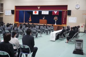 体育館の壇上に入学式、進級式と書かれた垂れ幕の下で壇上で話をされ、椅子に座り話を聞いてる生徒の様子を背後から撮影した写真