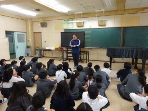緑色の黒板の前に、黒いグランドピアノが設置された教室内で、話をされる先生と向かい合わせで膝を抱えるように座り熱心に話を聞く生徒達を背後から撮影した写真