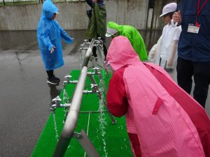 中央に設置された太いパイプから、いくつもの蛇口から水が排出されている様子を、ピンク色や水色、黄緑色の雨がっぱを着用した生徒達が熱心に見学している様子の写真