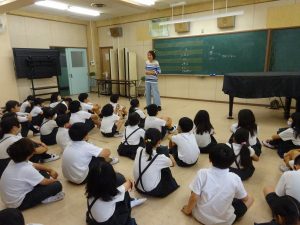 黒板前に黒いグランドピアノが設置された教室内で、前方に立ち話をする教諭と、向かい合わせで膝を抱えるように座り熱心に話を聞く生徒達を背後から撮影した写真