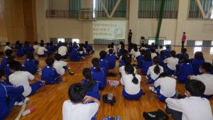 体育館の壁に投影されたペップトークを、白い体操服や青いジャージを着用した生徒達が、膝を抱えるように座りながら真剣に見ている様子を背後から撮影した写真