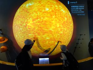 子どもたちの背丈よりも遥かに大きい画面に映し出されている太陽のような惑星に触れながら話をしている生徒2人の写真