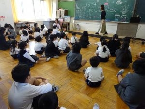幼稚園のお友達を歓迎するメッセージやイラストが描かれたカラフルな黒板を前に、教室内の床にたくさんの児童達が座り交流会を楽しんでいる様子の写真