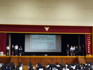 体育館内の壇上で、大きなスクリーンを使い遠足について学んだことや感想を投影し、スクリーン両側には4年生10名ほどが立ち発表している様子の写真