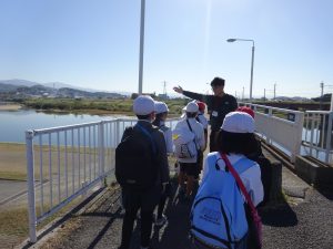 後方に大和川が望める白いフェンスが設置された場所で引率の先生が片手で大和川を指しながら説明をしておりその話を聞いている生徒たちの後ろ姿の写真