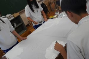 机いっぱいに広げられた白い団旗の前で数名の生徒たちが色を塗っている様子の写真