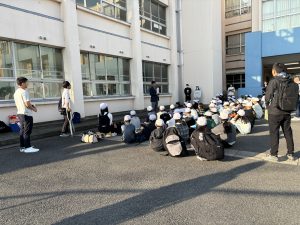 校舎の前で先生方が前に立ち話をしており、三角座りをしながら話を聞いている生徒たちの写真