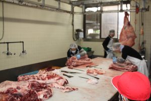 天井から吊るされた大きな牛肉を、従業員の手によってテーブルの上に部位ごとに切り分けられている様を、赤い帽子を被った生徒が見学している様子の写真