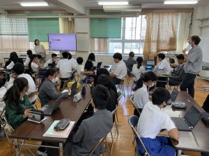 4名ずつ座る長机がある教室内の中央に、大きなモニターが設置され、女性の先生がモニターを表示させながら説明し、熱心に生徒達が聞いている様子の写真