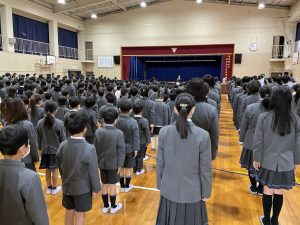 体育館内で、縦一列に整列した生徒達が壇上の先生の話を聞いている様子を背後から撮影した写真