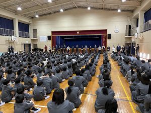 体育館内で、縦一列に整列し床に座った生徒達が壇上の前に横一列に並んだ先生を見ている様子を背後から撮影した写真