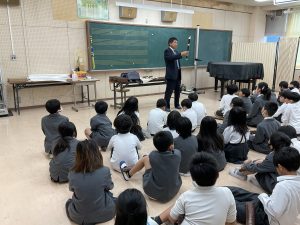 黒板の前に黒いグランドピアノが設置された教室内で、黒と白のリコーダーを右手に持ち話をされる先生と向かい合わせで膝を抱えるように座り熱心に話を聞く生徒達を背後から撮影した写真