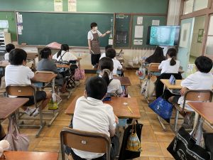 エプロンを着用しテレビモニターを使い説明する先生と、自席に座り熱心に話を聞く生徒達を背後から撮影した写真