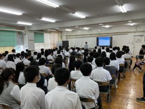 教室内に設置されたテレビモニター画面に、たくさんの生徒達が用意されたパイプ椅子に座り、非行防止動画を熱心に見ている様子を背後から撮影した写真