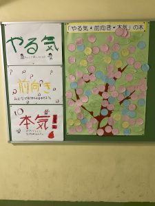 壁に掲示されている「やる気・前向き・本気」の木と文字が書かれ、メッセージの書かれたピンクや水色などの丸い紙が色とりどりの満開の木のように見立てた絵や生徒たちの制作物の写真