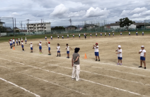 先生が見守る中、運動場のトラックを囲むように等間隔に生徒たちが立ち、学園エイサーの練習の様子の写真