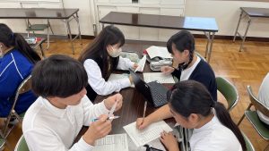 1つの長机に男女4名が並んで座り、パソコンを使い向かい合わせでプレゼンテーションしている様子の写真