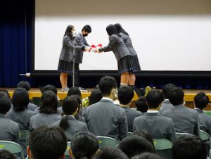 体育館の壇上の上で2人ずつ向かい合い、赤いリボンの付いたメッセージを手渡している写真