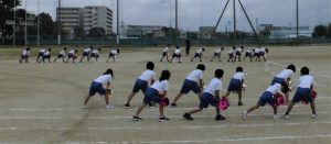 運動場で3グループほどに数名分かれて集まりそれぞれポンポンを持ち体勢を揃えながら体を動かしている練習の様子の写真