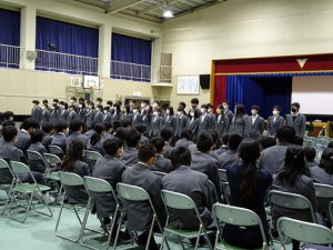 体育館に椅子に座った生徒が、前方で立って歌っている様子の生徒の歌を聴いている写真