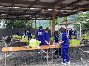 屋根のある屋外の調理スペースに長机が並んでおり、グループごとに分かれてテーブルで生徒たちが調理作業をしている様子を写した写真