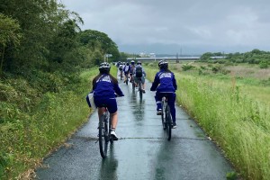 曇り空の中、両側が草の茂った堤防の道を、ヘルメットをかぶり自転車に乗ってツーリングを楽しんでいる生徒たちの後ろ姿を写した写真
