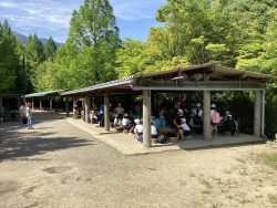 新緑に囲まれた自然豊かな屋外で、屋根付きで壁の無い建物に子どもたちが集まっている写真