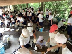周りに青々とした樹木が並んでいる場所で、子どもたちが協力しながら食べる準備をしている様子の写真