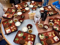 丸いテーブル上にハンバーグやサラダ、白ご飯、スープ、お茶などが器に盛り付けられて8名分並んでいる写真